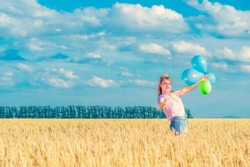 Naklejka na ściany i meble A young girl with colorful balloons in her hands is standing among a yellow wheat field against a blue cloudy sky. Looks into the camera.