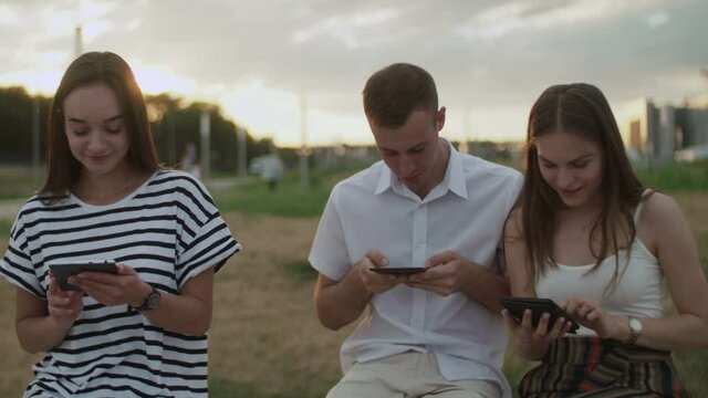 Two girls and a guy sitting in the park during the day reading an e-book
