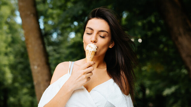Young indian brunette woman in white summer dress eating vanilla ice cream in waffle cone, with closed eyes enjoying taste of food, outdoors. Beautiful elegant girl on summer vacation in park