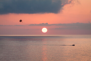 Parasailing behind a boat at sunset over the sea with the sun goes down. Silhouette of a person with parachute against evening sky with clouds. Beautiful seascape.