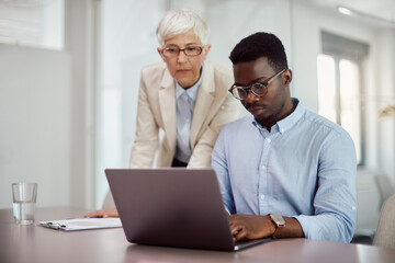 African American businessman works on laptop while mature mentor supervises him in office.