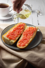 Toast with tomato, traditional Spanish breakfast. Olive oil is poured from a glass jug, side view. Pan tumaka