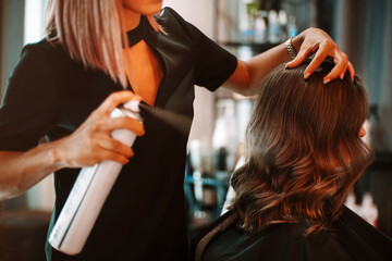 Female hairdresser fixing customer woman hair with spray and put care treatment while styling curls