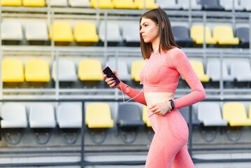 Young attractive fitness model listening music on smart phone charching positive energy before workout outside.