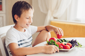 Boy sitting in the kitchen eating healthy vegetables cucumber and tomato