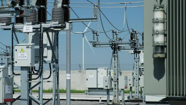 Windmill generates electricity and equipment of high voltage substation at switchyard on foreground against blue sky in summer