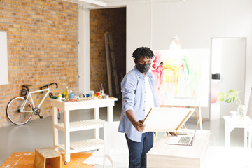 African american male painter wearing face mask looking at artwork in art studio