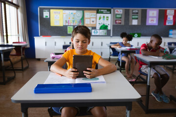 Caucasian boy using digital tablet while sitting on the desk in the class at elementary school