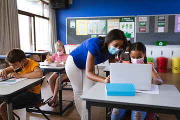 African american female teacher wearing face mask teaching a girl to use laptop at elementary school - Powered by Adobe