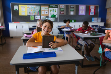 Portrait of smiling caucasian boy holding digital tablet sitting on the desk at elementary school