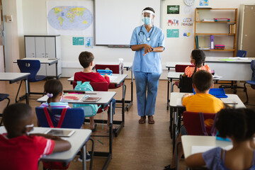 Female health worker wearing face shield talking to students in the class at elementary school