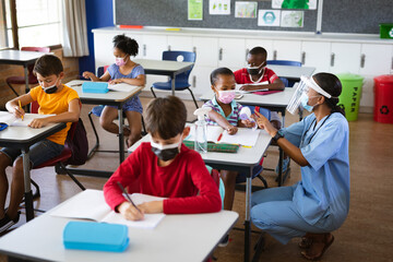 Female health worker wearing face shield measuring temperature of a girl at elementary school