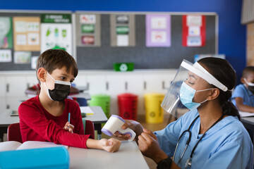Female health worker wearing face shield measuring temperature of a boy at elementary school