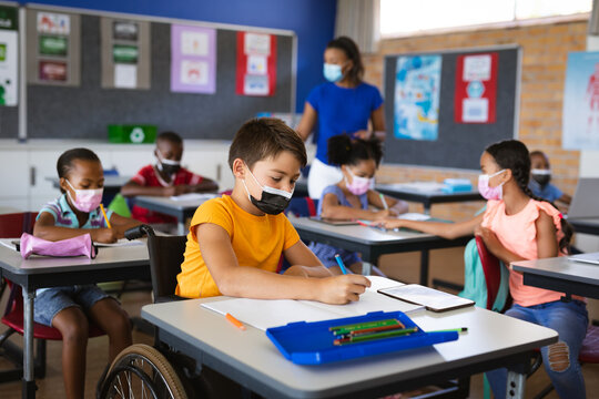 Disabled caucasian boy wearing face mask studying while sitting on wheelchair at elementary school