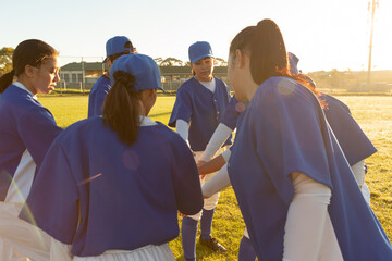 Diverse group of female baseball players warming up on field at sunrise, stacking hands
