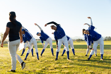 Diverse group of female baseball players with coach, warming up on field, stretching from the waist