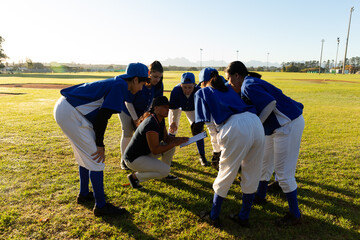 Diverse group of female baseball players standing in huddle around squatting coach on field