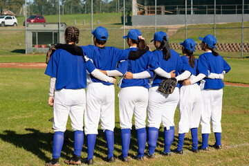 Diverse group of female baseball players standing on field with arms around each other before game