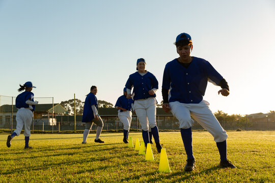 Diverse group of female baseball players warming up on field at sunrise, running slalom around cones