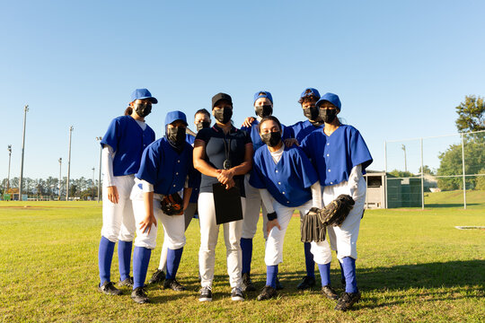 Portrait of diverse group of female baseball players and coach in face masks standing on sunny field - Powered by Adobe