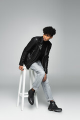 trendy african american man in jeans and leather jacket looking away on high stool on grey background