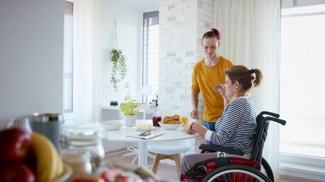 Disabled mature woman in wheelchair sitting at the table with a son indoors at home, eating.