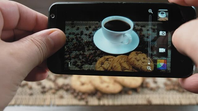 A man takes pictures of a cup of coffee and cookies on his cell phone. Coffee beans are also scattered on the table. A man takes photos of a still life on a table. He has a phone in his hands.
