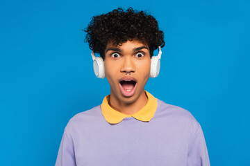 thrilled african american man in wireless headphones looking at camera isolated on blue