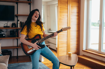 Young cheerful woman playing electrical guitar at home while dancing