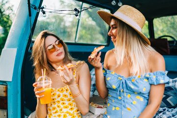 Two attractive cheerful women drinking lemonade near van and eating pizza, enjoying summer vibes in...