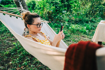 Young woman using mobile phone, relaxing in hammock outdoors, in the backyard garden.