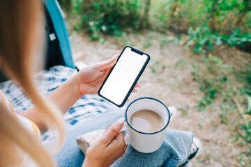 Woman holding a smartphone with a white screen mock up, sitting in a van and drinking tea during...
