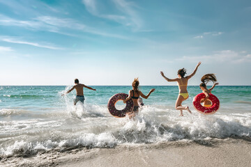 Happy cheerful friends resting on a beautiful beach at sunny day and running into the sea with inflatable rings. Tropical vacations concept
