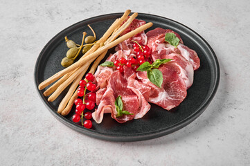 Close up prosciutto coppa di parma ham slices with basil, grissini sticks, capers and redcurrant on dark plate on textured background