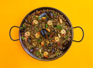 a traditional spanish black paella with shrimps, mussels and squid ink in a pan on yellow...