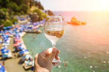 Glass of wine in hand. Glass with young fresh white wine on the background of the Mediterranean beach and the sea in a tourist town in the summer under sunlight. Summer, travel, lifestyle, relaxation