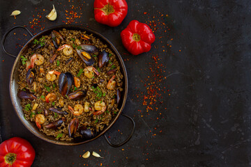 spanish black paella with shrimps, mussels and squid ink in a pan with tomatoes and chili paper on a black background. view from above, flat lay, Copy space