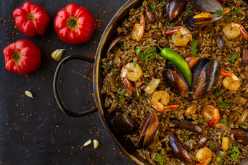 Close up of a traditional spanish black paella with shrimps, mussels and squid ink in a pan with tomatoes on a black background. view from above, flat lay
