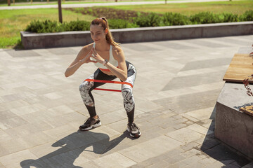 Woman in tracksuit does squats with elastic bend in city park