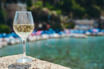  Glass of white wine against the background of the Mediterranean beach and the sea in a tourist town in the summer under sunlight. Summer, travel, lifestyle, relaxation, and enjoyment concept.
