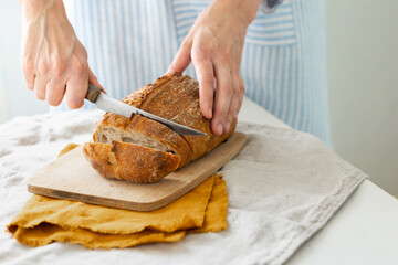 women's hands hold bread against the background of a light apron, a woman cuts bread, bread with a crispy crust is beautiful and appetizing, delicious bread lies on a wooden cutting board and a