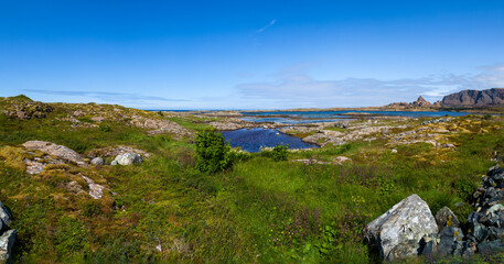 Fototapeta premium Panorama of the rocky ocean coast on Leka island overlooking the mountains and Heilhornet peak on the horizon under blue clear skies. Trondelag county, Norway