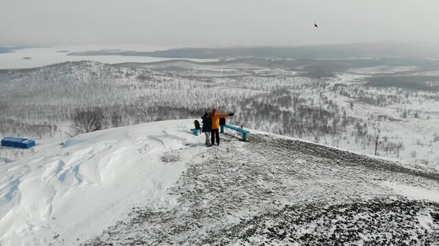Man and woman are standing on top of snow-covered hill. They stopped to rest. They wave their hands. You can see snow slopes, trees and factory chimneys. Smoke is coming from the pipes. Aerial view