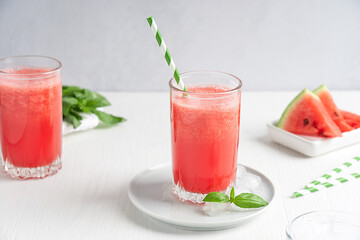 Refreshing cold juice or smoothie made of red ripe watermelon served in drinking glass with paper straw and basil leaves on white wooden background with fruit slices at kitchen in hot summer season