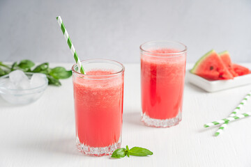 Watermelon refreshing cold red juice served in two drinking glasses with paper straw and green basil leaves on white wooden background with fruit slices as dessert and ice cubes summer season.