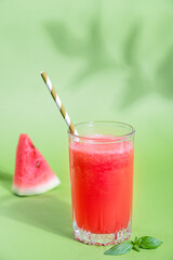 Cold refreshing red watermelon juice in drinking glass with paper straw and basil leaf served with slice of raw fruit under summer sunlight with plant shadow against green background. Vertical image