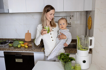 Young caucasian mother with blonde hair hugging baby daughter eight months old and drinking detox cocktail in glass with fresh green vegetables on kitchen at home