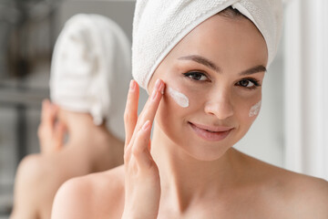 Close up cropped portrait of a young beautiful woman in bath towel smiling applying face body cream...