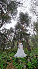 A bride in a white dress stands at the trees in the mountains