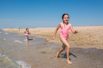 Happy kids girls running on sandy beach. Children enjoying and having fun at the sea on sunny day. Family summer holidays and summertime concept.